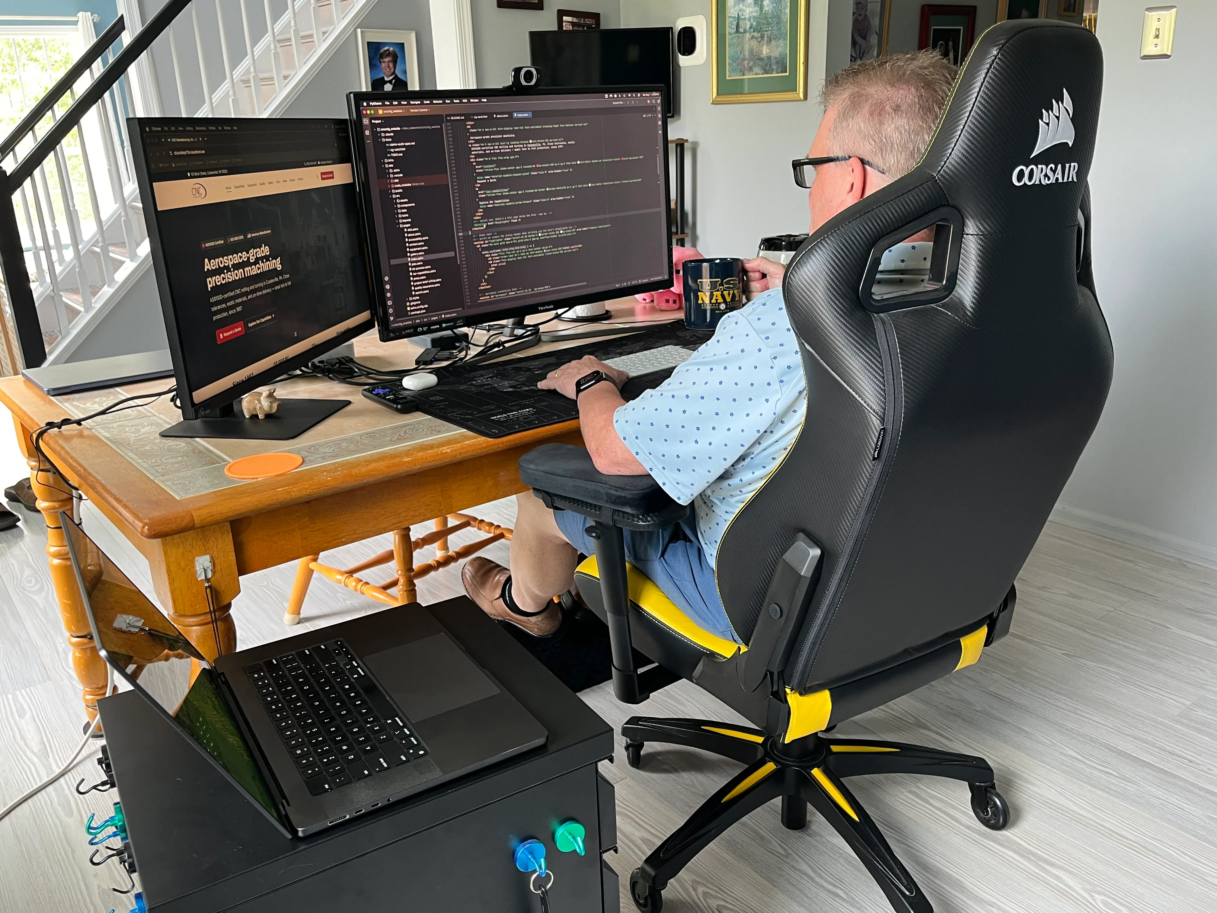 Bill at the keyboard in a black-and-yellow racing-style chair, coffee mug in hand, with the new CNC Manufacturing homepage — “Aerospace-grade precision machining” — on one monitor and its code open in the editor on the other