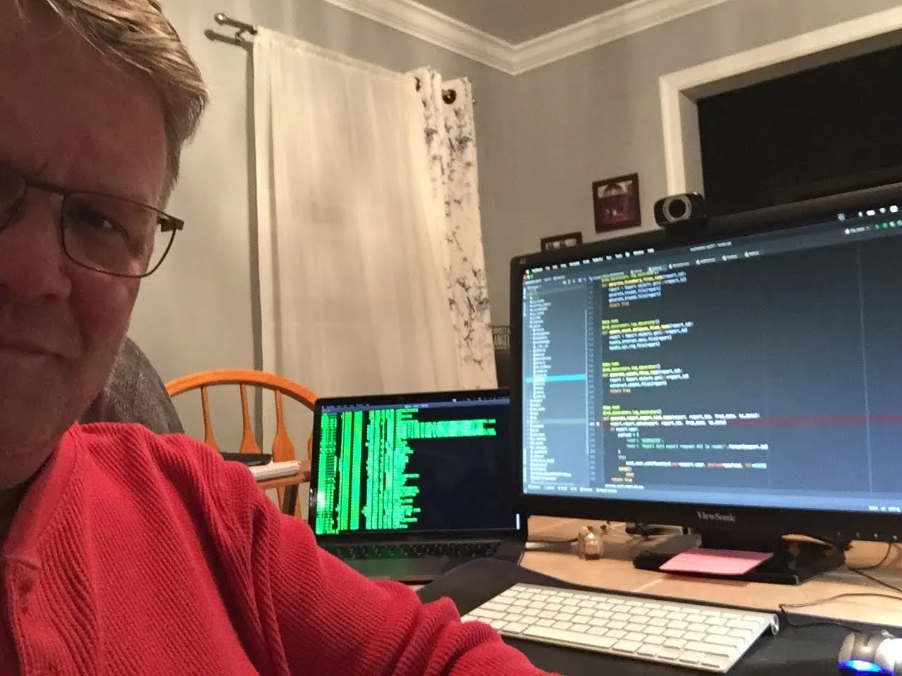 Bill leaning into the frame at night in a red hoodie and glasses, half-smiling, while a laptop scrolls green logs and a big ViewSonic glows with code