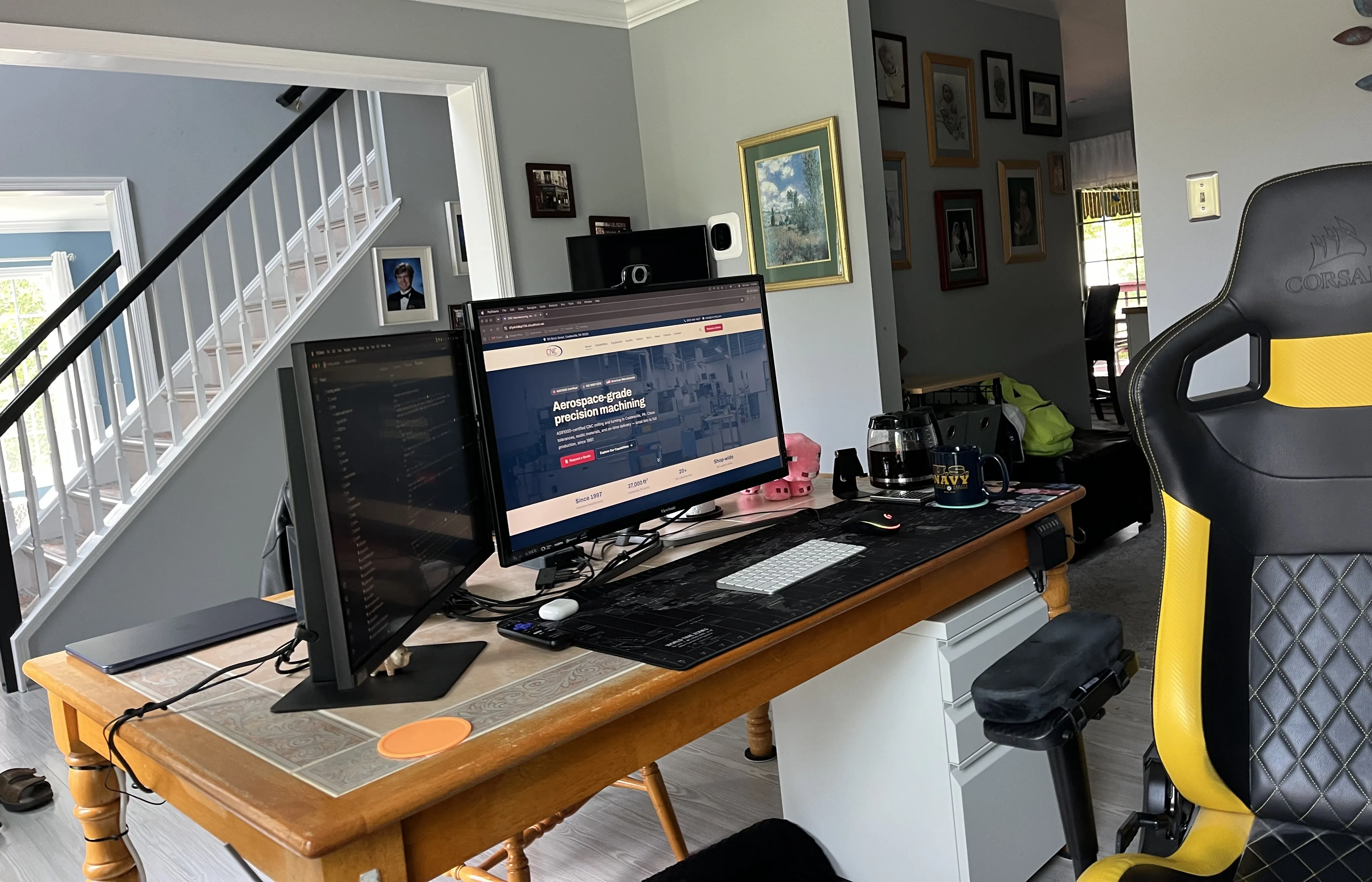 Bill’s current dual-monitor setup on a tile-topped kitchen table, the CNC Manufacturing site on one screen and an editor on the other, with the racing-style chair pulled up, a full pot of coffee standing by, and a calculator and small plush pig on the tidy desk