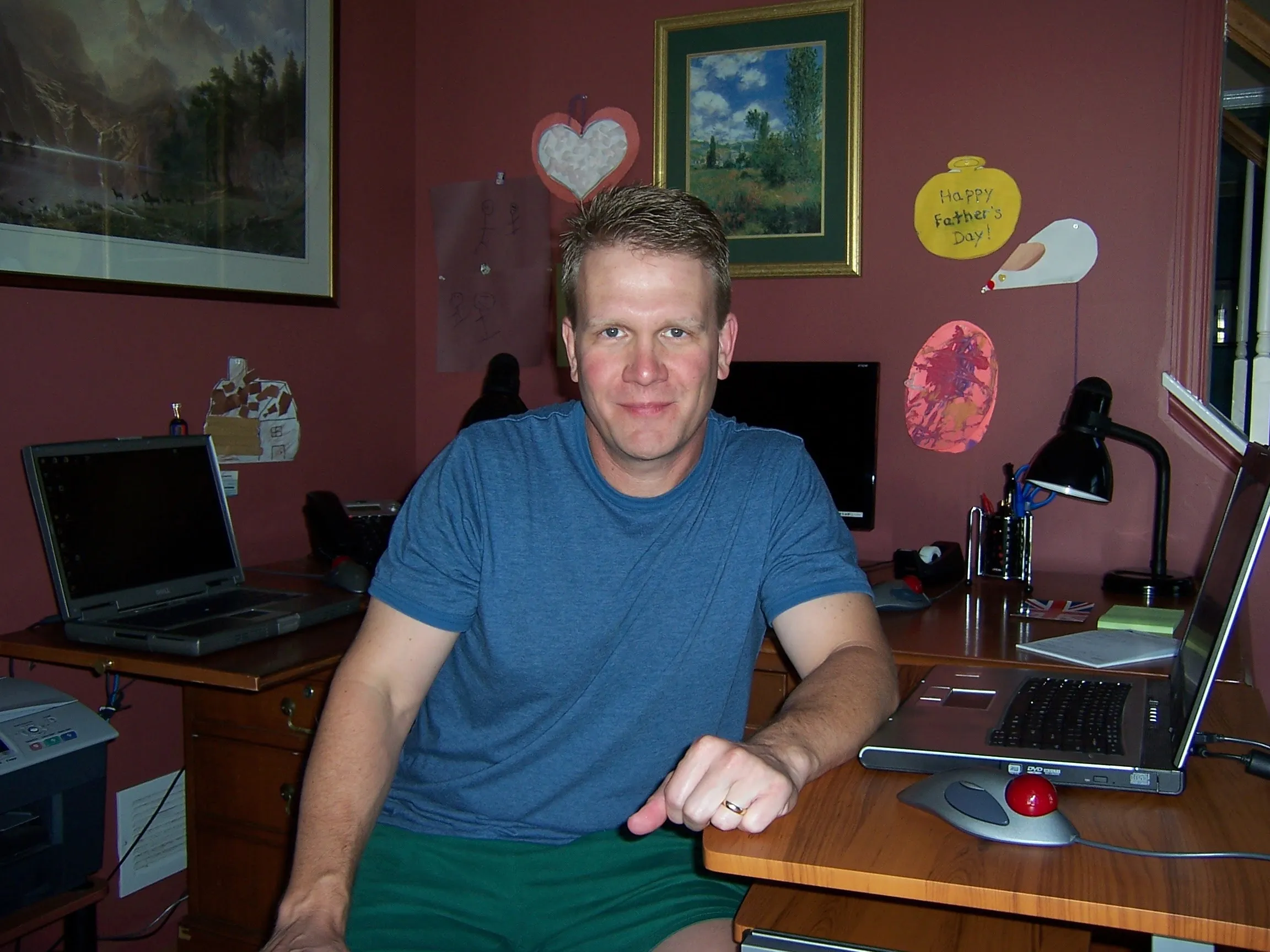 A younger Bill in a blue tee perched between two open laptops, a desktop glowing in the corner and a kid’s “Happy Father’s Day” drawing taped to the wall