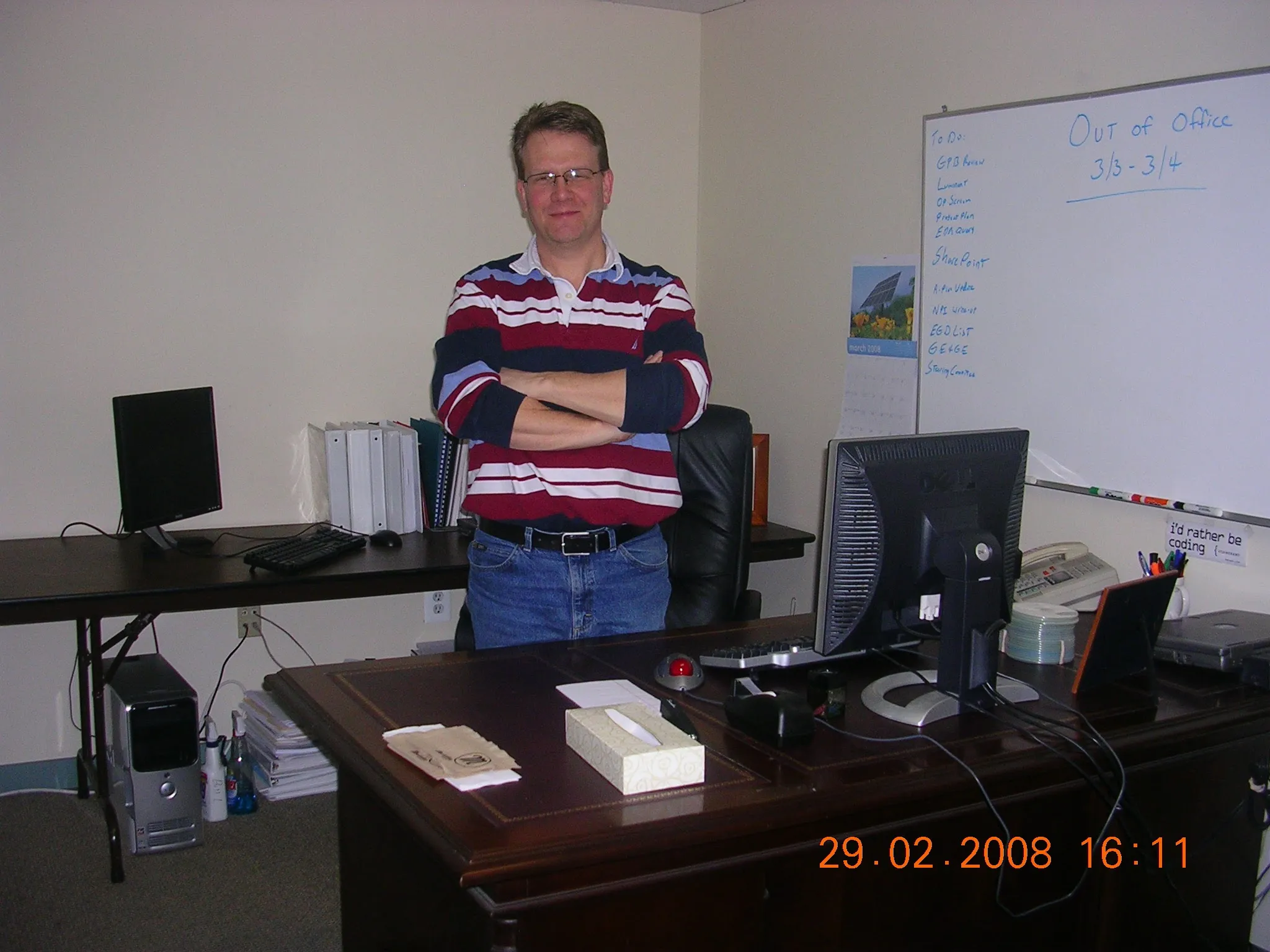 Bill in 2008, arms crossed and looking faintly pleased with himself in a striped sweater, between two Dell setups and a whiteboard that reads “Out of Office”