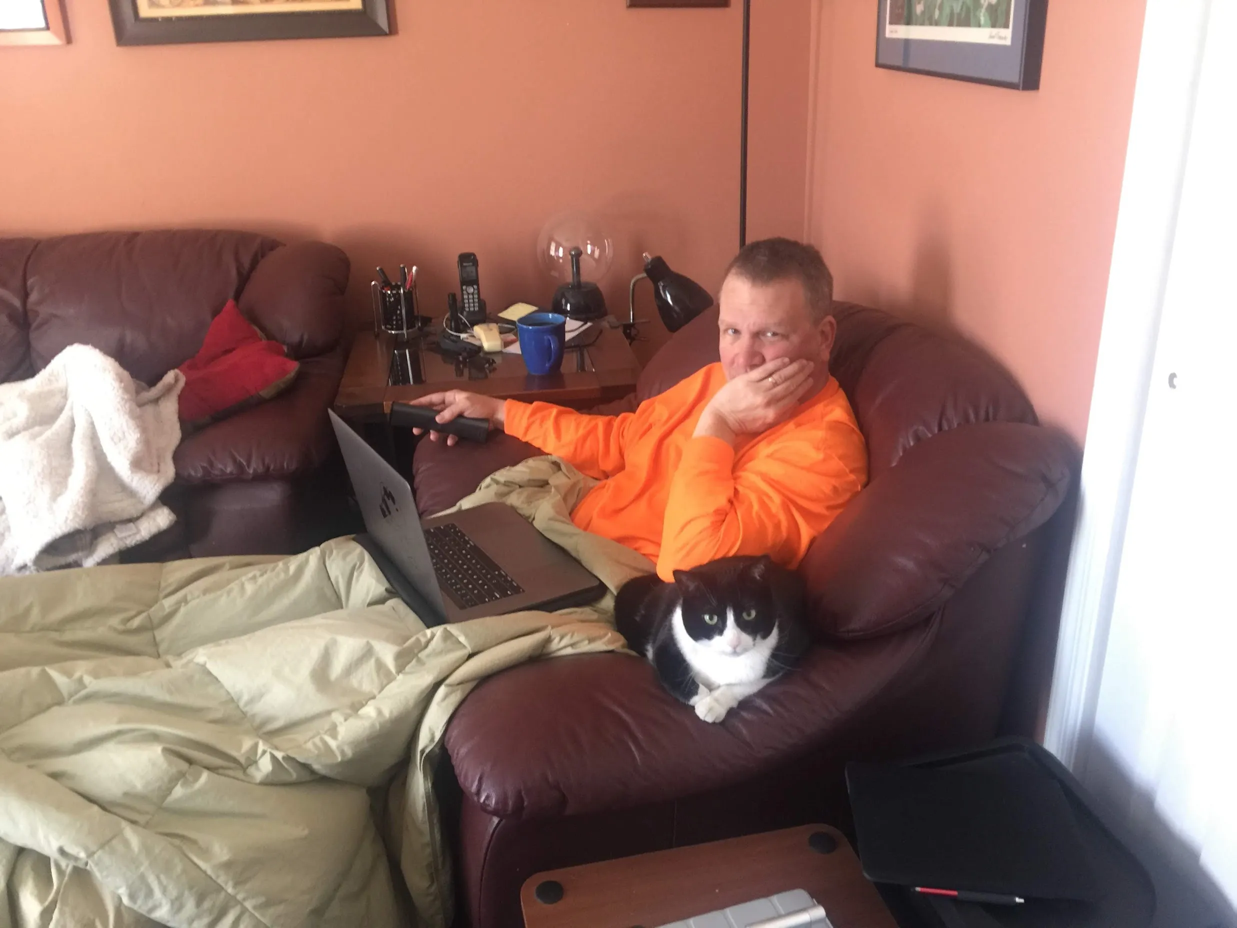 Bill reclining in a bright orange shirt with a laptop on his lap, a black-and-white cat planted firmly on the armrest beside him